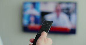 Woman hand switches TV remote control with finger, viewing of news, TV programs, modern technology for controlling equipment at a distance. Blurred TV in the background. Close up view.