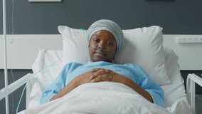 Zoom in shot of young African American female patient with nasal cannula and headwrap lying on hospital bed with hands clasped on her stomach and posing for camera with smile - Powered by Shutterstock - Get 15% off with code: PIKWIZARD15