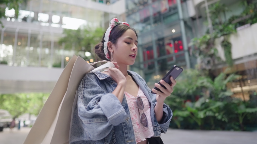 Attractive Smiling asian young adult woman shopper customer standing inside city modern department store holding shopping bags, using smart phone mobile for online navigator, promotion point checking  Royalty-Free Stock Footage #1091096929