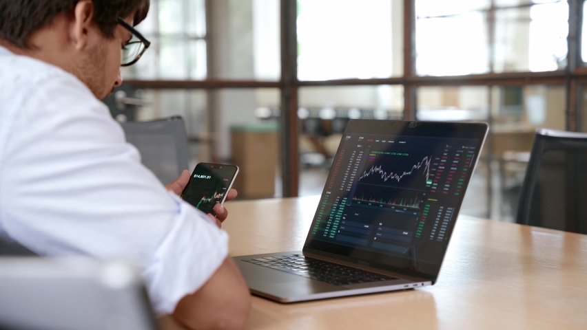 Indian business man trader broker investor analyst analyzing stock exchange market crypto trading chart data, managing financial risks looking at laptop using mobile app on smartphone. | Shutterstock HD Video #1091097059