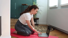 Online Yoga. Sporty young brunette caucasian woman turning on video lesson on laptop and start meditation practice, relaxed sitting in lotus position with closed eyes indoors at modern home interior