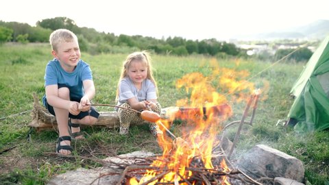 Brother and sister have conversation and cheerfully smiling while they roasting a  sausages on the sticks over the campfire flame in tent camp. Happy family or outdoor picnic activities concept image.