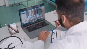 Videocall chat with female older patient and indian male doctor in modern clinic hospital using pc laptop computer, consulting online remotely. Telemedicine healthcare concept. Over shoulder view.