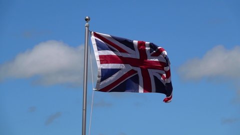Slow motion video of a Union Jack Flag fluttering in the wind with a blue sky and clouds background