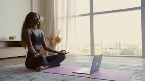 Young calm fit healthy African woman sitting on floor at home doing yoga breathing exercise, meditating learning online training virtual class on computer. Exercises for mental health concept.