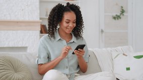 Smiling African American teen girl holding smart phone chatting in social media, watching video content, texting messages, playing games, using mobile apps, surfing internet on sofa at home.