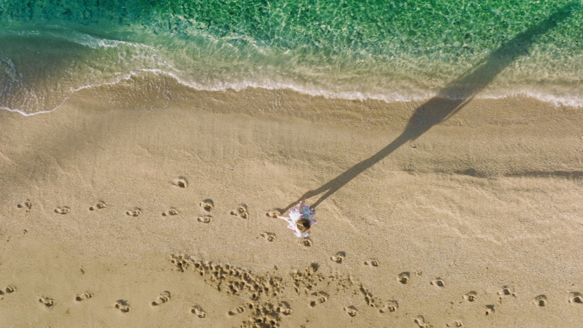 Aerial overhead shot alone woman walking by empty sandy beach. Long person shadow on pure beach with teal green clear sea waves running to the shore. Summer sunrise background, scenic Hawaii nature 4K