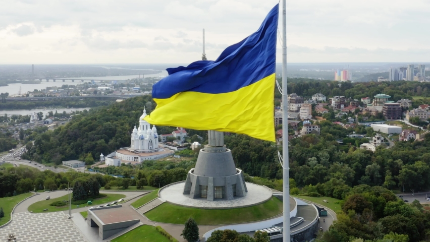 Aerial top view of Kiev Motherland statue monument and national ukrainian flag fluttering on foreground in Kyiv, Ukraine