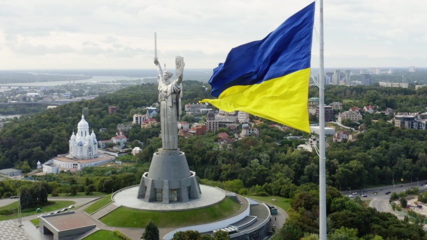 Aerial top view of Kiev Motherland statue monument and national ukrainian flag fluttering on foreground in Kyiv, Ukraine