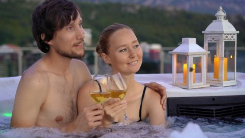 A young man and woman are relaxing in the hot tub on a rooftop with a view on mountains during sunset