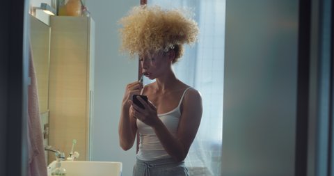 Cinematic authentic shot of young happy smiling brazilian woman is using technology smartphone for work or entertainment while brushing her teeth with toothpaste in bathroom at home.