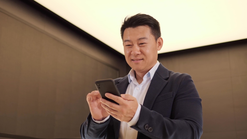 Confident korean man using mobile phone app in modern light office. Positive asian business guy browsing financial news smiling reading good news standing indoor. Successful male chinese adult.  Royalty-Free Stock Footage #1091121601