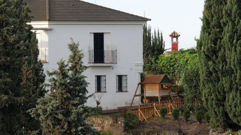 Two-storey white facade house with security bars on the windows and small wooden playhouse for kids in the yard. Quiet place with scenic high pine trees near the building. Cordoba in Andalusia, Spain