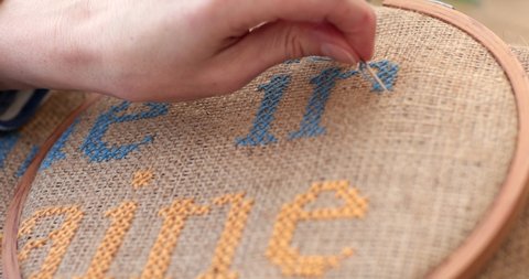 POV hand hold needle and embroider cross stitch letter n with blue thread on burlap fabric in embroidery hoop. Yellow embroidered text - Ukraine. Traditional needlework in blue yellow colors on canvas