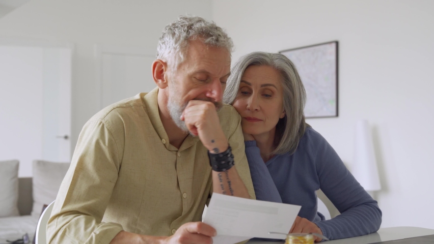 Older mature couple checking bank documents using laptop at home. Senior mid age retired man and woman reading paper bills, calculating pension or taxes, planning retirement finances, doing paperwork.