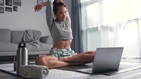 Sporty young Asian woman exercising at home, watching fitness video on Internet or having online fitness class, using laptop, living room interior.