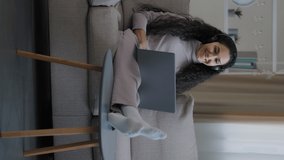 Vertical view happy young arab woman girl with curly hairstyle sit on cozy sofa talk at headset microphone use video call broadcast greet friend online communicate in social networks use wi-fi at home