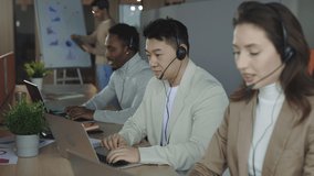 Serious asian male worker sitting at table with laptop and using headset to give advice to clients. Multiracial group of call center employees talking with customers in modern office. - Powered by Shutterstock - Get 15% off with code: PIKWIZARD15
