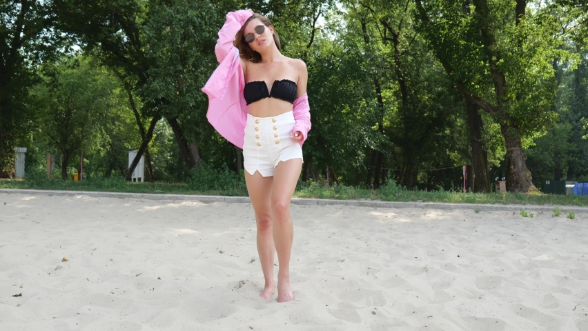A young woman in a black bra and white shorts stands on the sand. A woman in a pink shirt. slow motion