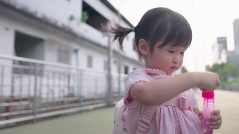 3 years old Asian girl with backpack playing with camera by blowing soap bubbles after parents pick her up from elementary school, kid playtime, healthy child, life insurance, asian girl blow bubbles 