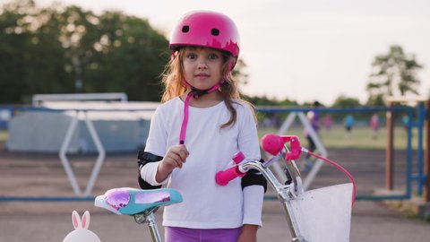 Portrait of yong beautiful caucasian light hair girl. She leans her elbow on the bike seat and looks into the camera. Kid wears pink helmet and elbow pads. Sport activity for children concept.: stockvideo