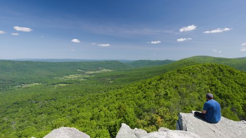 Rockland, WV - USA - May 30 2022: A bearded man wearing a hat and sunglasses sits on a rock ledge at Big Schloss overlooking the Trout Run Valley on a beautiful summer day. Static, wide-angle shot.