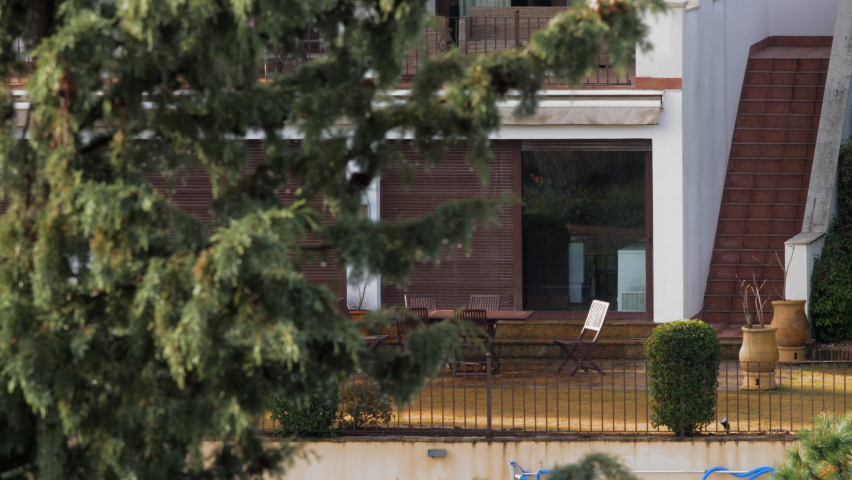 Detail of house exterior. Empty table and chairs in yard by half shuttered window and side stairway leading to upper floor. Pine tree branch in foreground, sunny morning. Cordoba in Andalusia, Spain