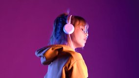 Gen z teen girl model wear headphones listening dj pop music playlist dancing on purple violet neon light color studio background. Stylish 20s hipster fashion woman in hoodie sunglasses at cool party.