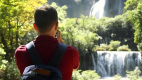 Happy male tourist with backpack in travel. Wanderlust, tourism concept. Young man traveler make vertical video for social media of amazing waterfalls and tropical nature on his smartphone camera.