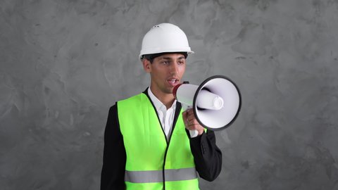 An Arab man in a white helmet and vest stands on a gray background and reads an announcement through a bullhorn.