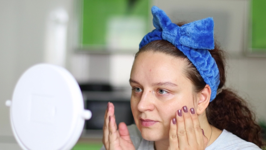 Curly woman in hairband applying and smearing moisturizing lotion or skincare cream for skin lifting. Looking in mirror, face self massage. Circle and patting movements, home spa procedures treatment