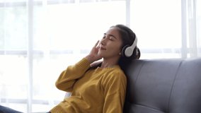 Asian woman listen song through wireless headphones, sit on sofa at home enjoy favourite music use digital streaming services, spend leisure use tech, hobby concept.