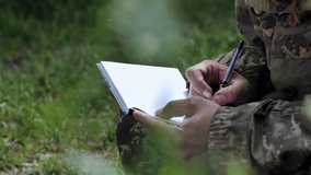 A soldier writes a letter while sitting on the grass. A serviceman of the Armed Forces of Ukraine makes notes in a notebook. Close-up of a pen in the hand of a military officer who writes in notebook - Powered by Shutterstock - Get 15% off with code: PIKWIZARD15