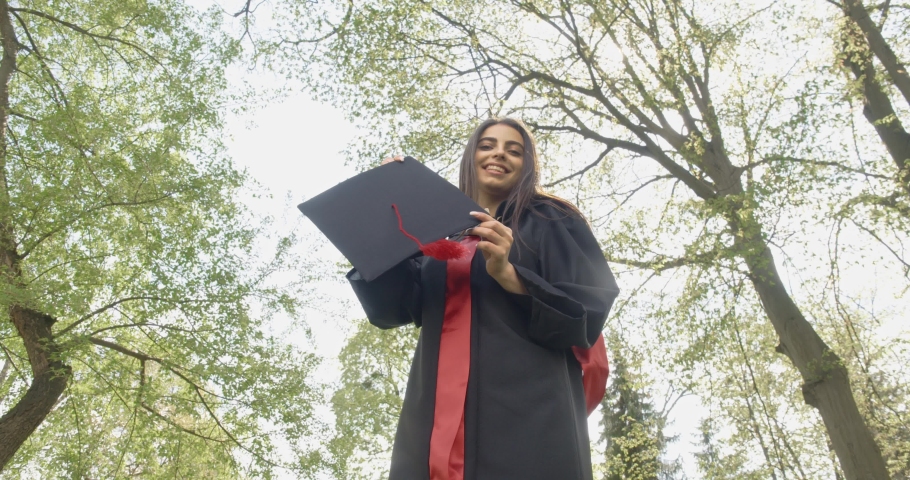 Front view of attractive female graduate standing in park, looking at camera, smiling, happy, glad. Brunette girl with long hair showing mortarboard. Concept of graduation.