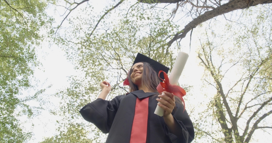 Close up of cheerful young lady putting off mortarboard, throwing up, jumping outdoors in park. Brunette female getting master, bachelor degree, glad. Concept of graduating.