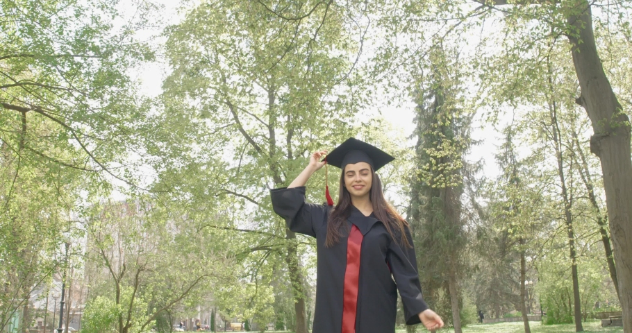Front view of pretty brunette girl getting off mortarboard, jumping, throwing up in park. Beautiful young female wearing graduation gown, smiling, cheerful. Concept of gradution.