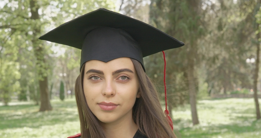 Front view of pretty girl wearing mortarboard and graduate gown standing in park, holding, showing diploma. Beautiful female looking at camera, smiling. Concept of education. 