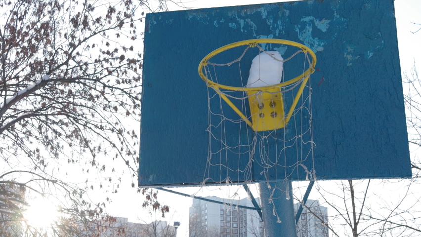 Close-up of an old blue basketball backboard with a yellow hoop, and a basket of threads swaying in the wind against the sky and tree branches
