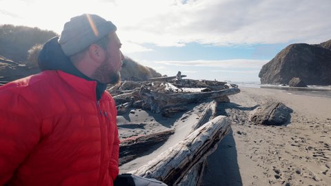 Lonely traveler in bright orange windbreaker exploring coastline with rocky formations behind. Close-up view of caucasian man enjoying beautiful panoramic ocean views. High quality 4k footage