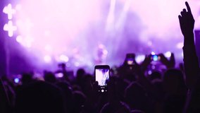 Music fan record video on phone in crowd on concert, closeup of audience people fans raise hands enjoy live music festival event shoot rock band stage on mobile device in purple lights, slow motion