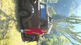Stylish woman in red dress and hat walking near classic vintage car. Young attractive modern woman walking in park near old truck and cactus on background. Vertical video