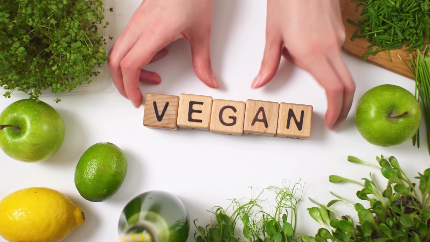 Girls put forward inscription vegan with their hands, assembled from wooden cubes. Green vegan ingredients with microgreens, sprouts, green apples, lemon, lime, green smoothie. Vegan food concept