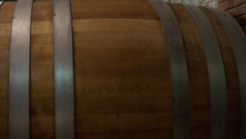 Close-up view of a wooden cask bound by metal hoops and a cellar master pouring red wine into glasses of visitors, pan left shot.