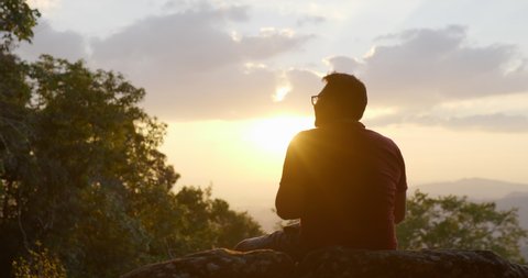 Slow motion in backlit shot, Young backpacker man sitting on rocky cliffs use digital camera take a photo while s in forest