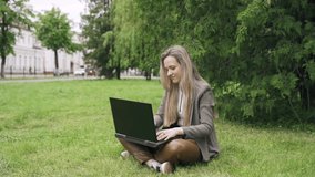 A young woman is sitting on the grass and typing on a laptop. Business and education concept.