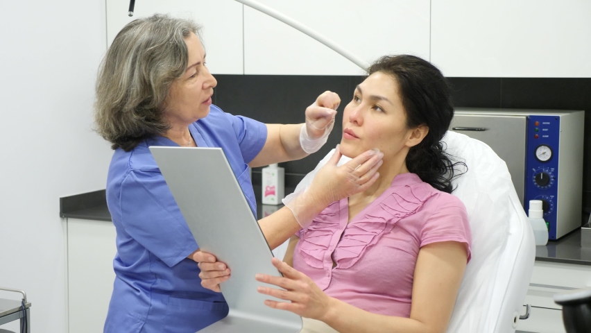 Adult asian woman looking at mirror while experienced female cosmetologist explaining her future hardware facial procedure in medical esthetic office Royalty-Free Stock Footage #1091199351