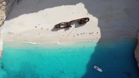 Aerial drone top view of the Navagio beach (Shipwreck beach) on the Ionian Sea coast of Zakynthos, Greece. Rocky cliffs, moored and floating boats, resting people, blue water.