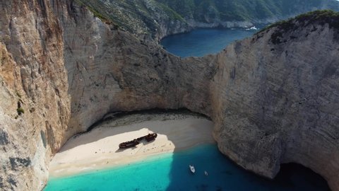 Aerial drone view of the Navagio beach (Shipwreck beach) on the Ionian Sea coast of Zakynthos, Greece. Rocky cliffs, moored and floating boats, resting people, blue water.