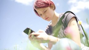 Young teen girl with dyed violet hair browsing internet using smartphone sitting on the green grass meadow while outdoor mountain walk. Active people, modern technologies 4K concept video