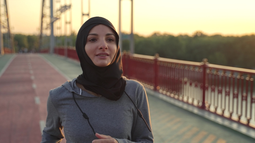 Close-up of pretty sporty arab woman wearing hijab running across footbridge early in the morning. Fit arab jogger enjoying morning cardio training on fresh air, healthy sports lifestyle concept - Powered by Shutterstock - Get 15% off with code: PIKWIZARD15